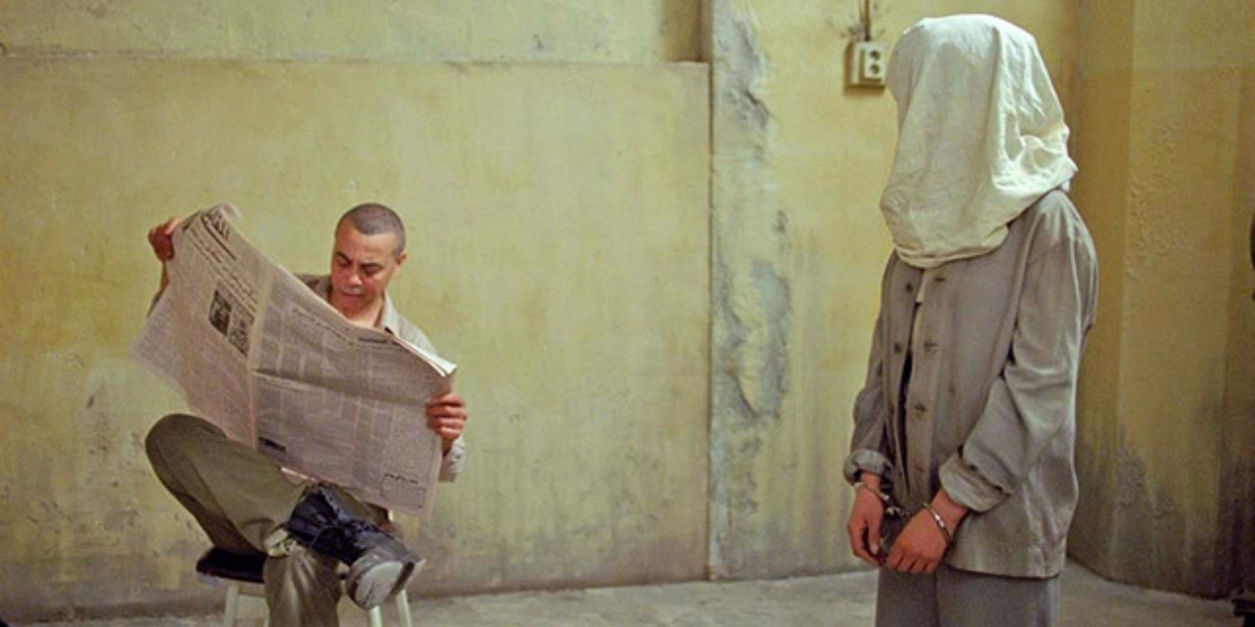 A man reads a newspaper while a woman stands besife it, with a bag covering her head