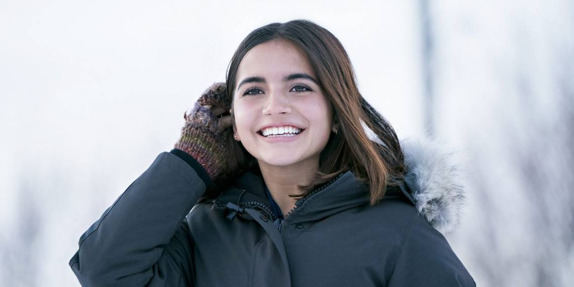 Aza smiles upward on a snowy winter's day. She wear mittens and a large coat and reaches up to put her hand behind her ear.