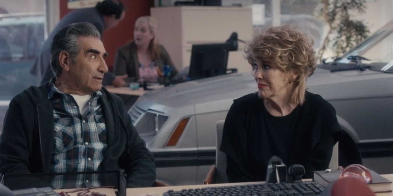 Johnny and Moira sitting at the desk at the car dealership
