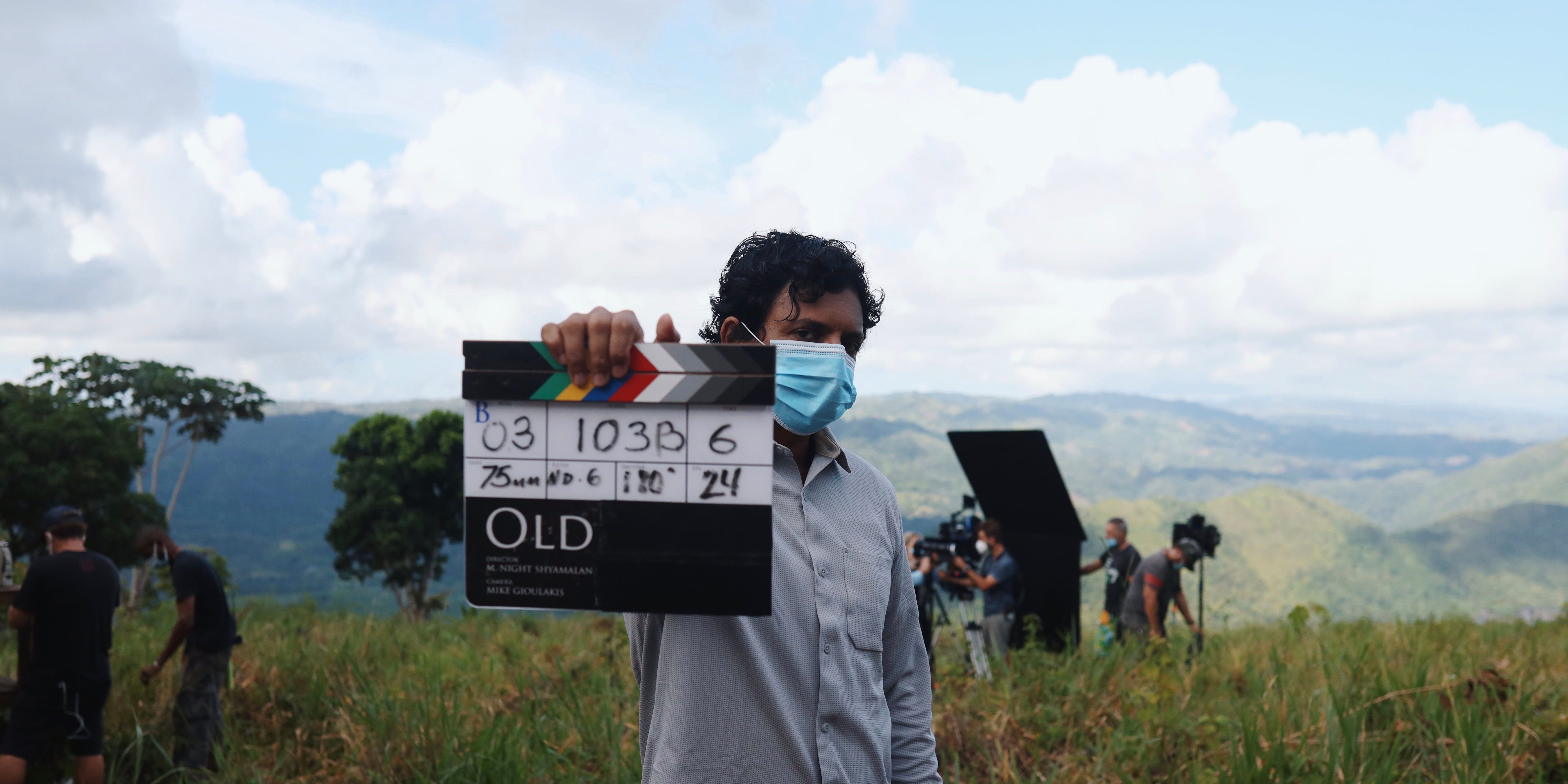M. Night Shyamalan holding up a clapperboard while working on his movie &quot;Old&quot; wearing a face mask