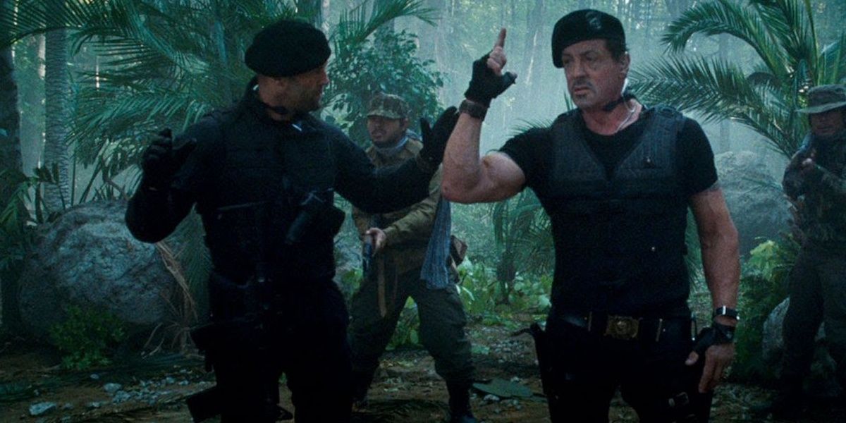 Sylvester Stallone signalling a sniper while Jason Statham stands with his arms raised, a still from The Expendables 2 