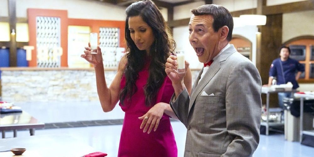 Paul Reubens in a grey suit holding a glass standing next to Padma Laxmi in a pink dress, also holding a glass, on Top Chef