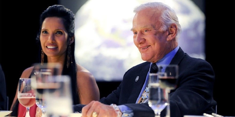 Buzz Aldrin in a black blazer and blue shirt sitting next to Padma Laxmi with a moon in the background