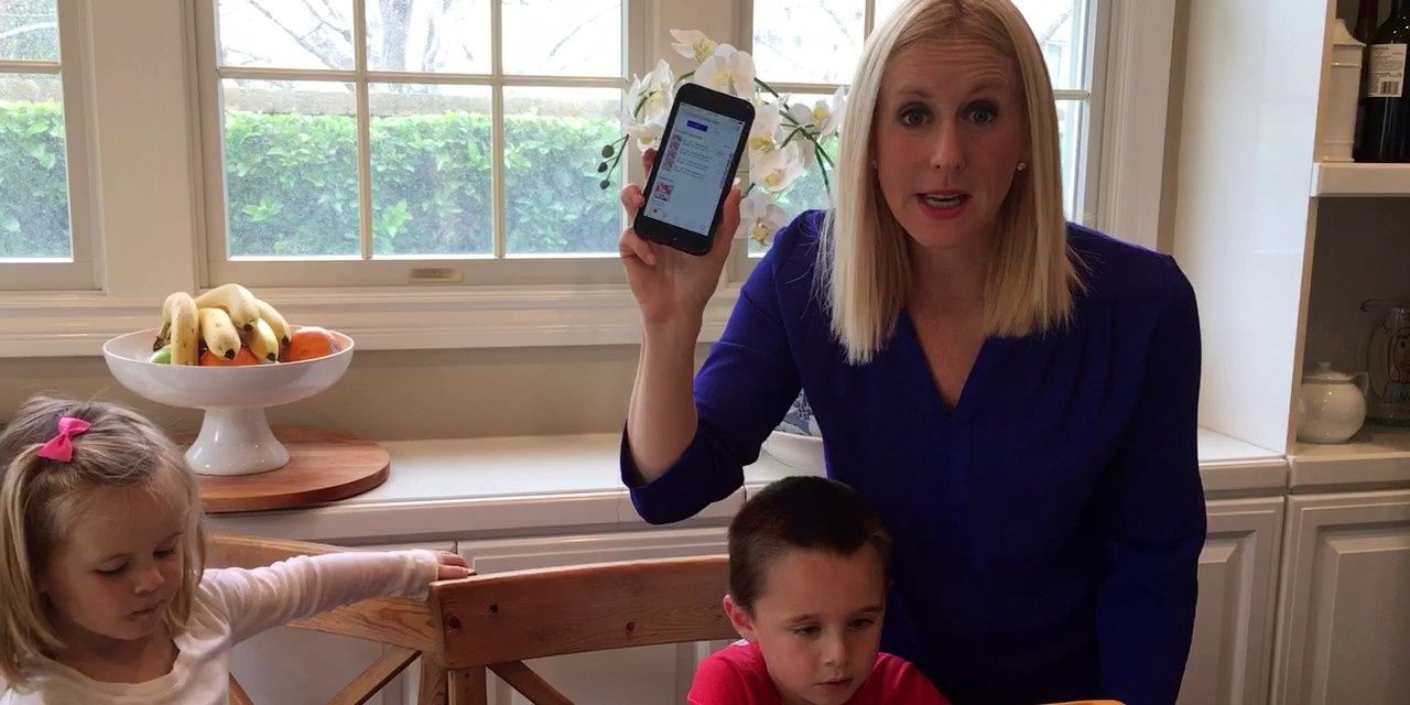 Kate Casey in a blue top holding up her phone with her children next to her sitting in a dining room