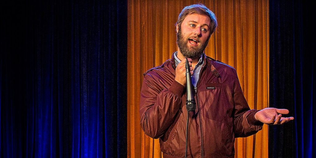 Comedian Rory Scovel in a brown leather jacket talking to the audience at his Netflix comedy special