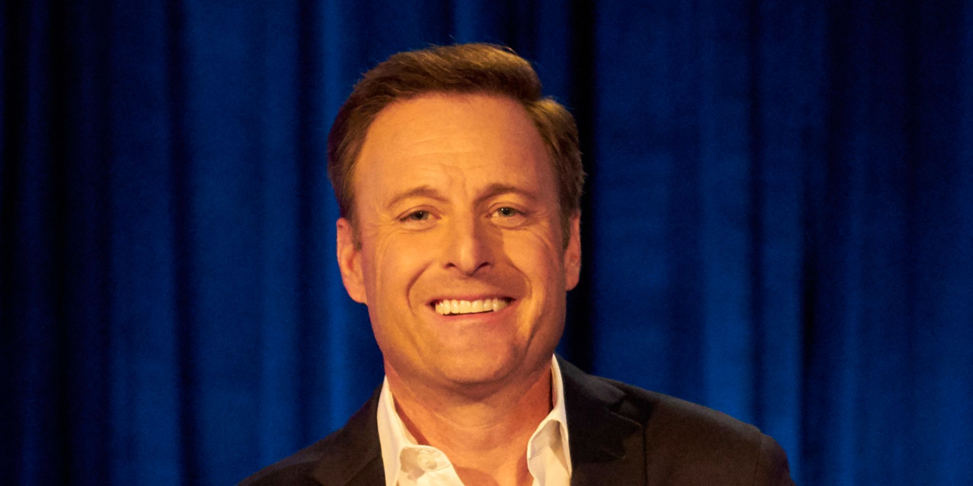 Chris Harrison smiling in front of blue curtain backdrop in The Bachelor