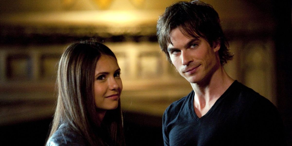 Elena smiles while standing next to Damon as they both look on at someone in the room.
