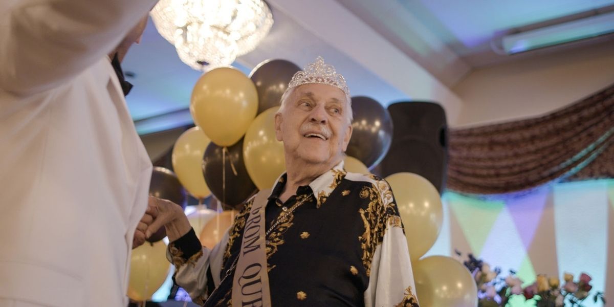 An elderly man wearing a Prom Queen crown dances