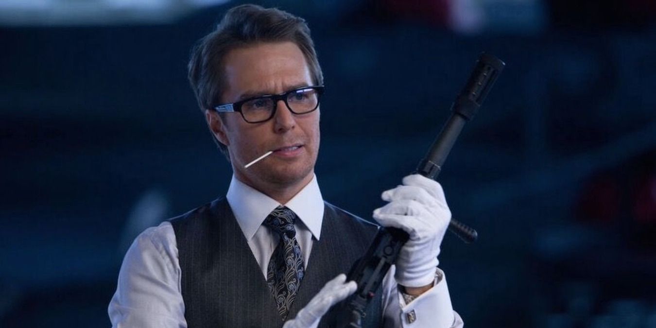 Justin Hammer holding a gun in the hanger, wearing gloves and a suit vest