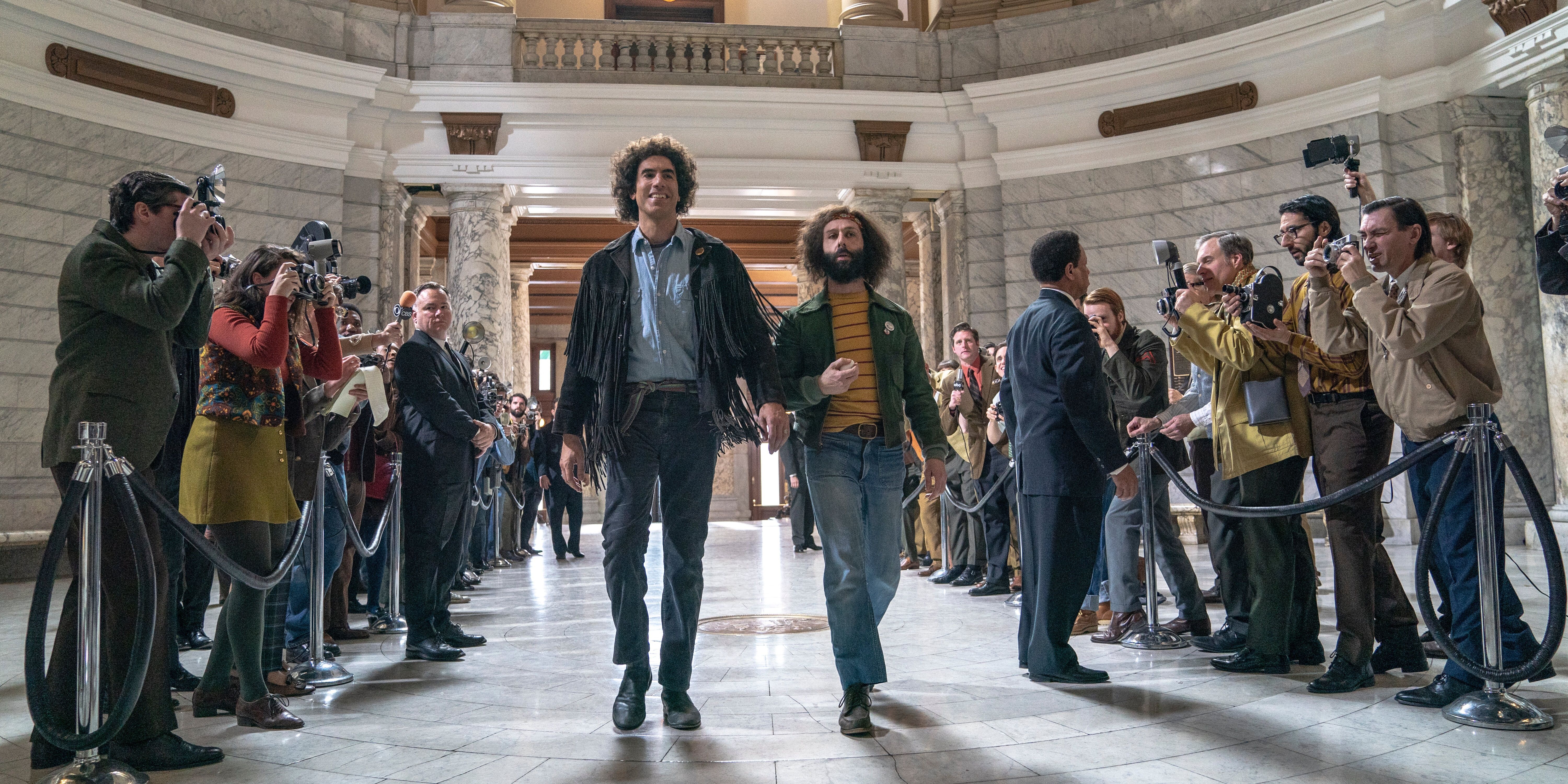 Sacha Baron Cohen and Jeremy Strong in The Trial of the Chicago 7 walking surrounded by reporters