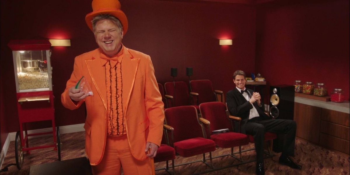 A blind man wearing an orange tux talks to the camera, while Daniel Tosh sits behind and laughs
