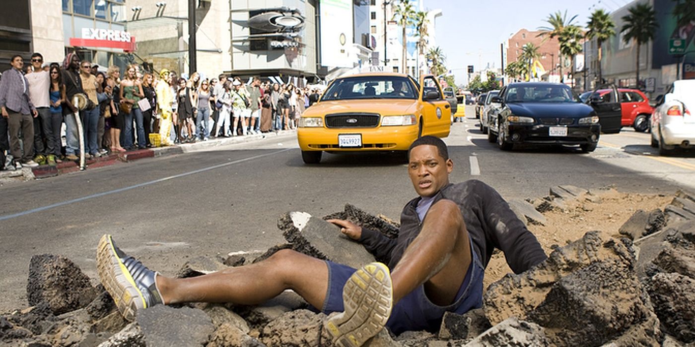 Will Smith laying on a destroyed road as passers by look on in a still from Hancock Cropped