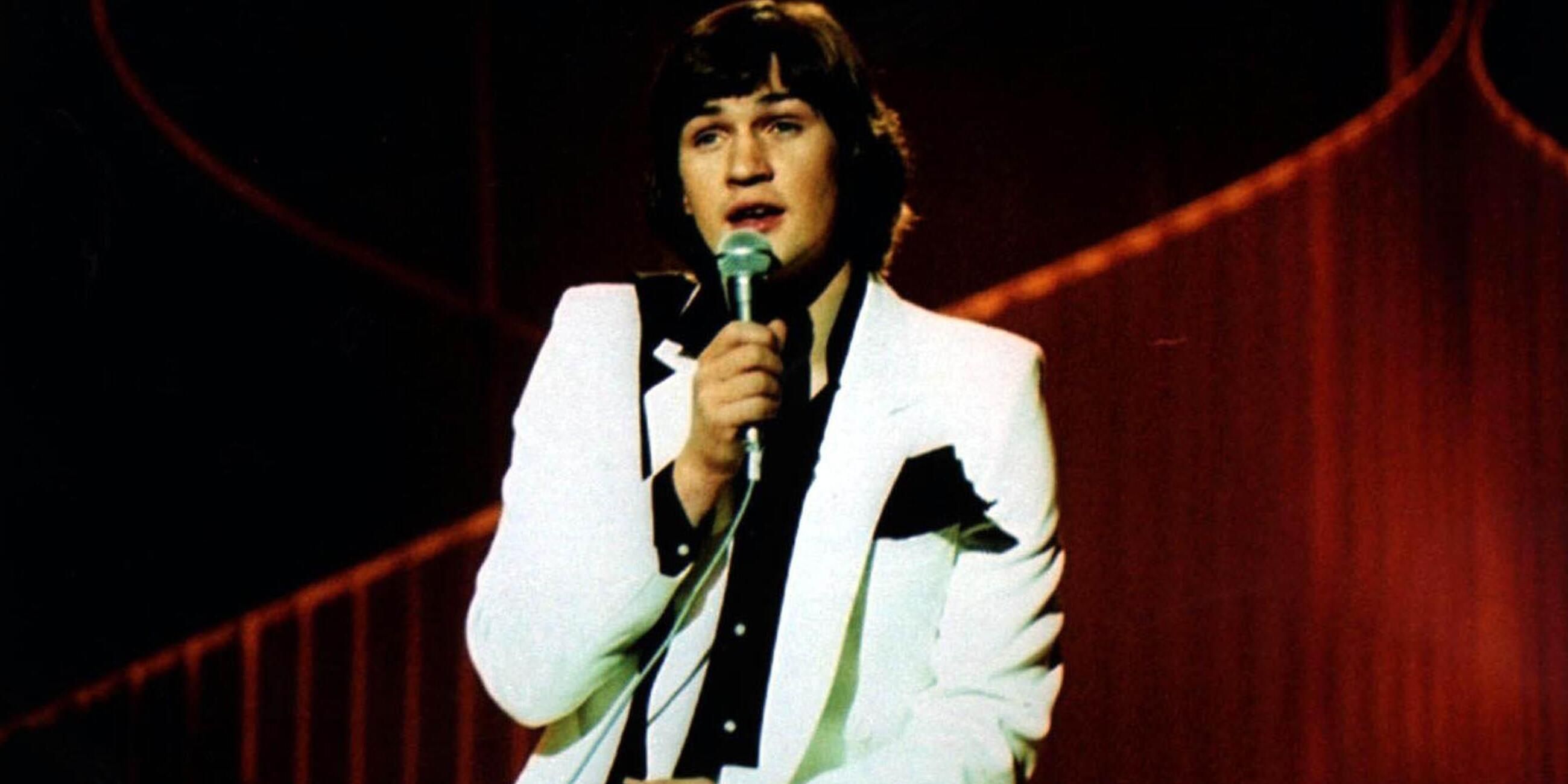 Johnny Logan holds a microphone on stage