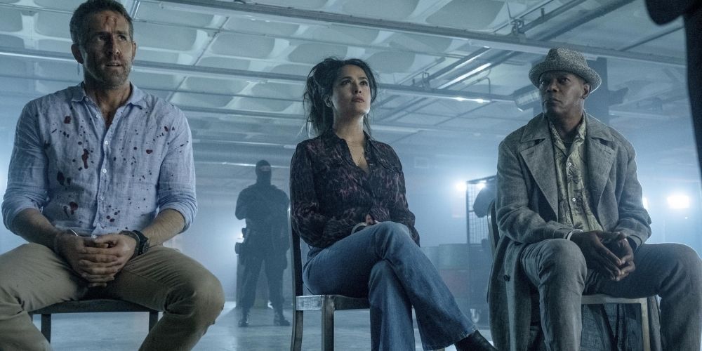 Kincaid, Sonia, and Bryce sit on chairs in a warehouse in The Hitman's Wife's Bodyguard