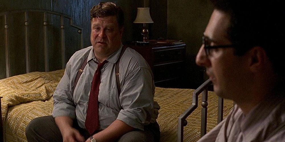 Charlie sits on the bed while talking to Barton at Barton Fink.