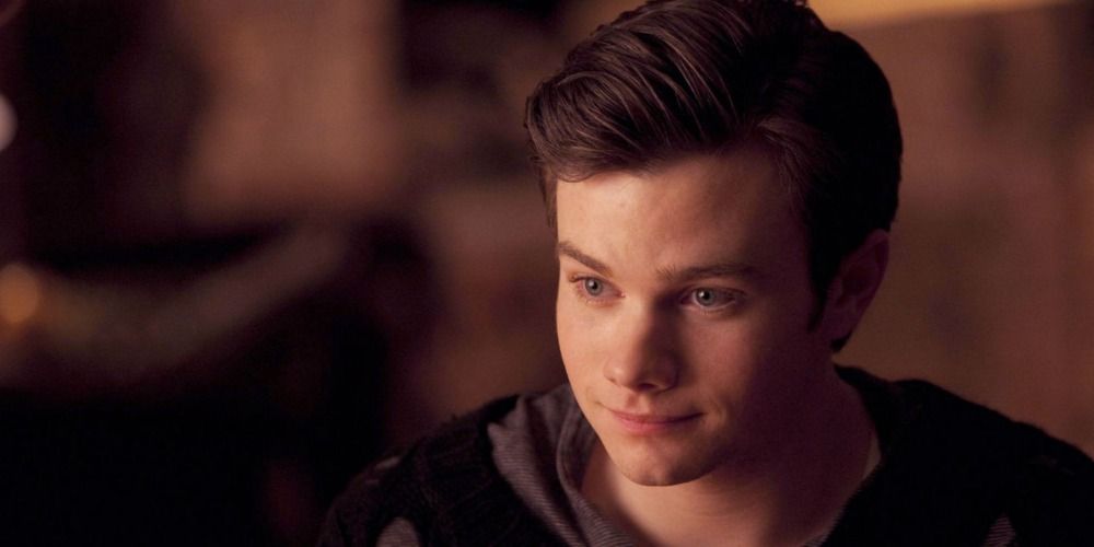 A young Chris Colfer in Glee smiling and looking off camera with a blurred background