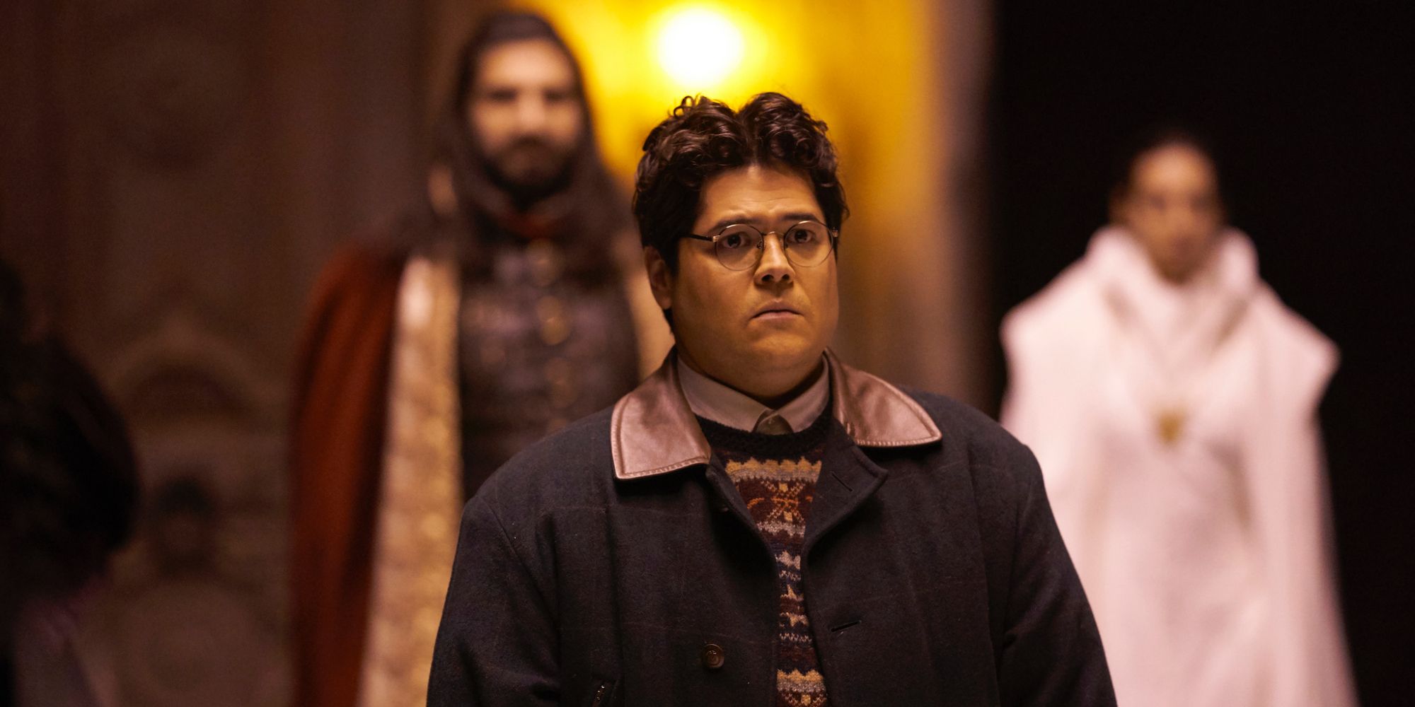 Guillermo stands looking sad as the vampires stand behind him in What We Do in the Shadows.