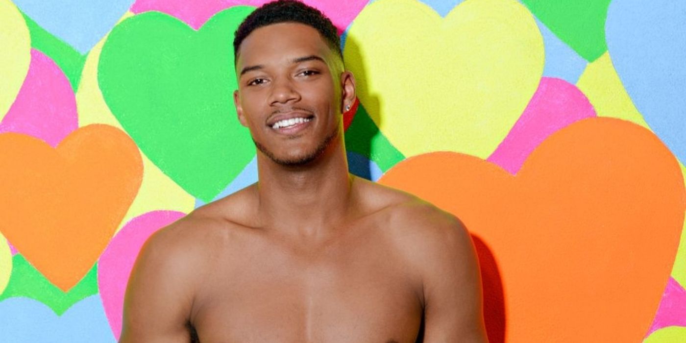 Theo Campbell smiling while standing against a wall full of multi-colored hearts in Love Island