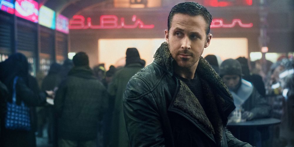 K wears a high collard jacket in front of neon signs in Blade Runner 2049.