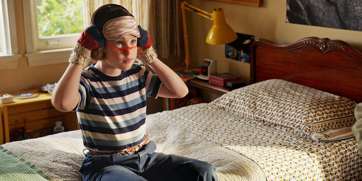 Sheldon sitting on the bed with tape around his face, headphones on, his hands up to his ears on Young Sheldon