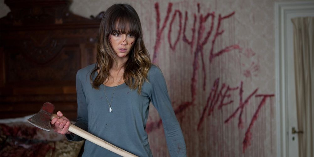Erin holds up ax with blood scrawled on the wall behind her in You're Next