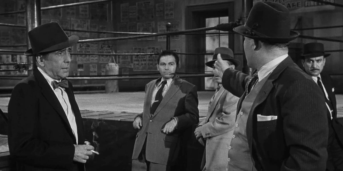 Humphrey Bogart looks at a man pointing a gun in a boxing ring in The Harder They Fall.