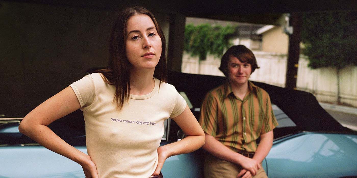 Alana Haim looks at the camera while Cooper Hoffman leans against a car from Licorice Pizza 