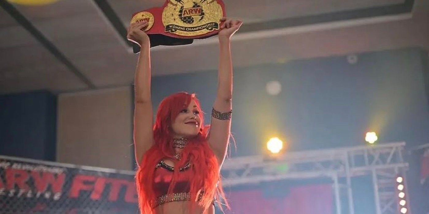 Paola Mayfield in her wrestling persona of Paola Blaze lifts a wrestling title belt over her head.