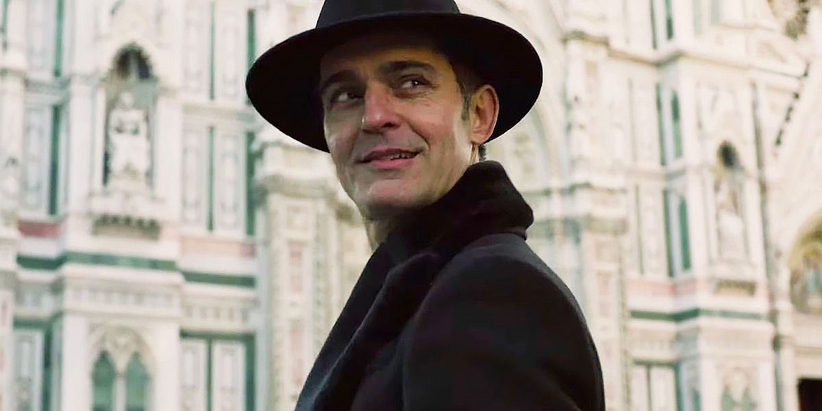 Berlin wearing a hat and a black coat as he stands in front of a church in Money Heist.