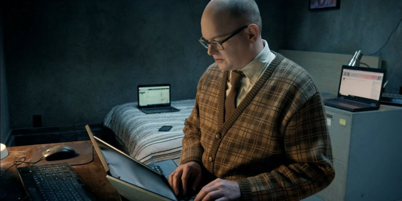 Colin wearing his plaid sweater as he uses a laptop in his basement room in What We Do In The Shadows.