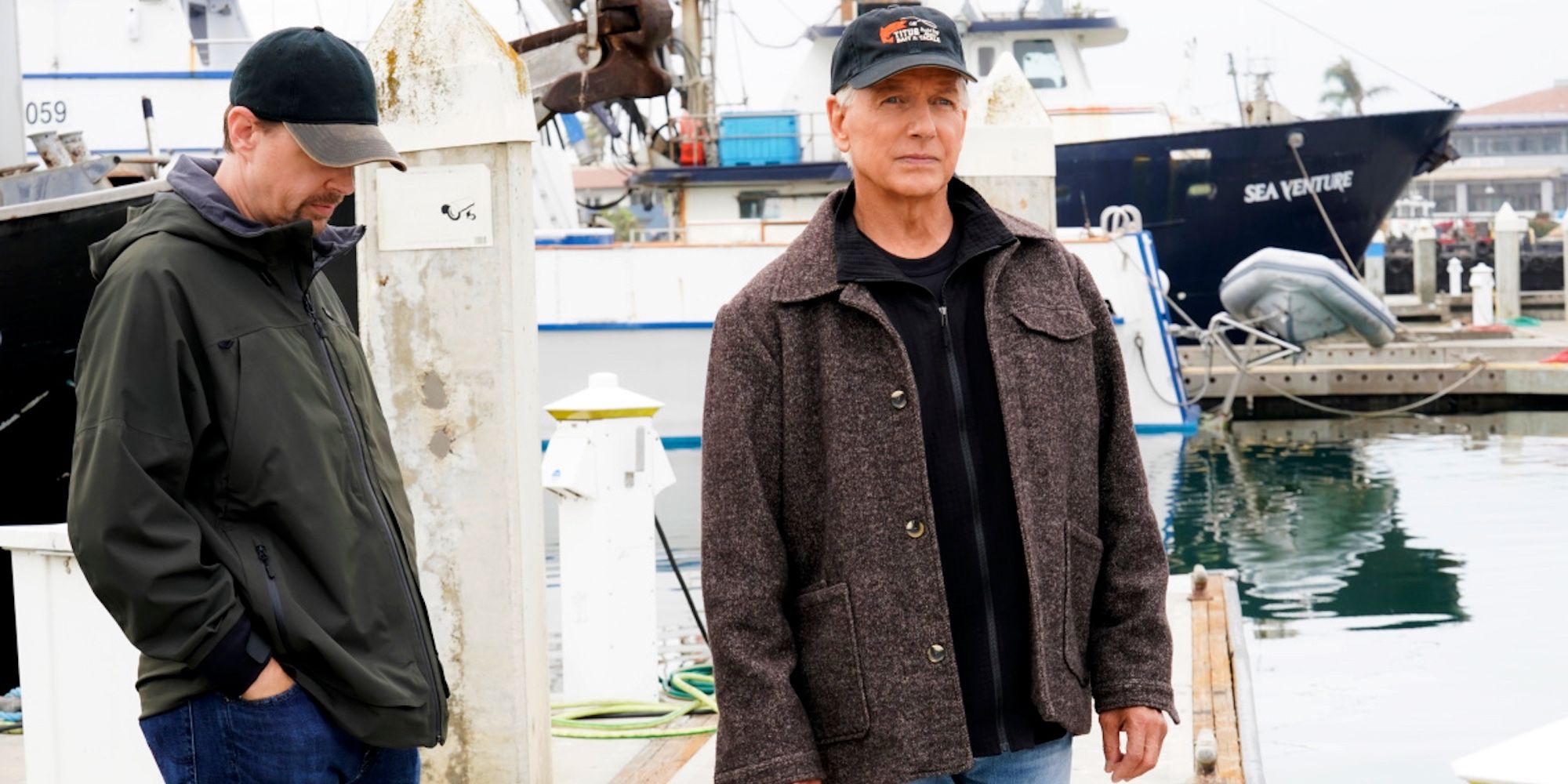 McGee standing behind Gibbs while they both wear jackets and caps at a marina in NCIS
