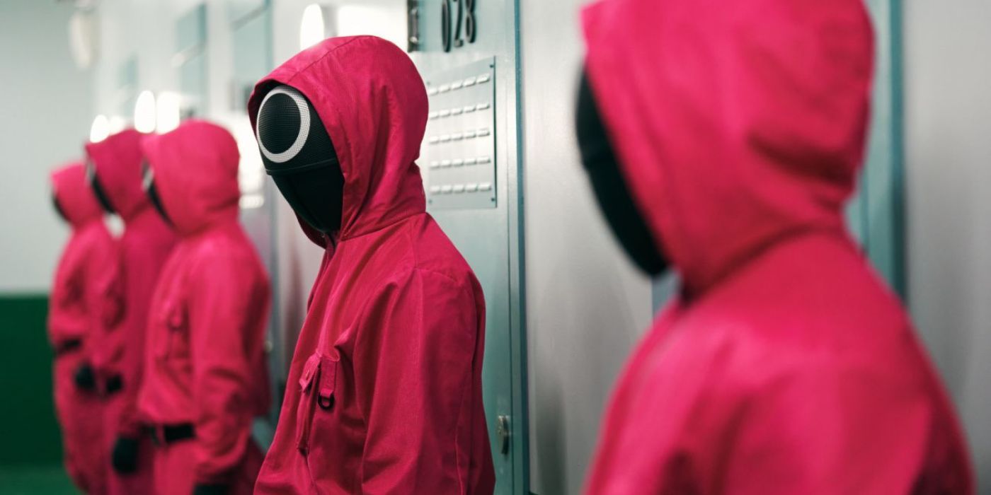 A guard wearing a pink jumpsuit and a black mask with a circle on it looks at the guard to his right in Squid Game.