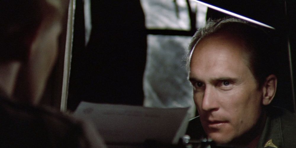 Robert Duvall in M.A.S.H. looks at someone off camera in the shadows.