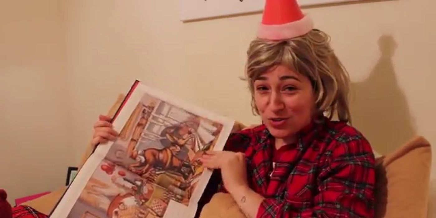 Melissa Villasenor holding a Christmas book and wearing a Santa hat with a blonde wig acting as Owen Wilson in a sketch from Saturday Night Live.