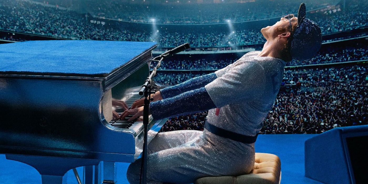 Taron Egerton as Elton John, wearing a baseball uniform playing the piano and leaning back singing in front of a crowded audience in a scene from Rocketman.