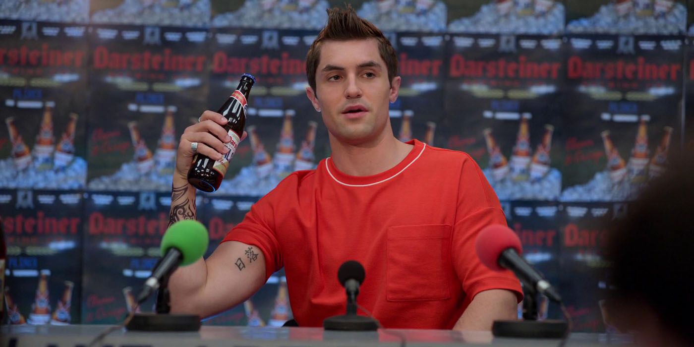 Jamie Tartt from Ted Lasso wearing a red T-shirt with a mohawk, holding a bottle of beet with a mic in front of him.