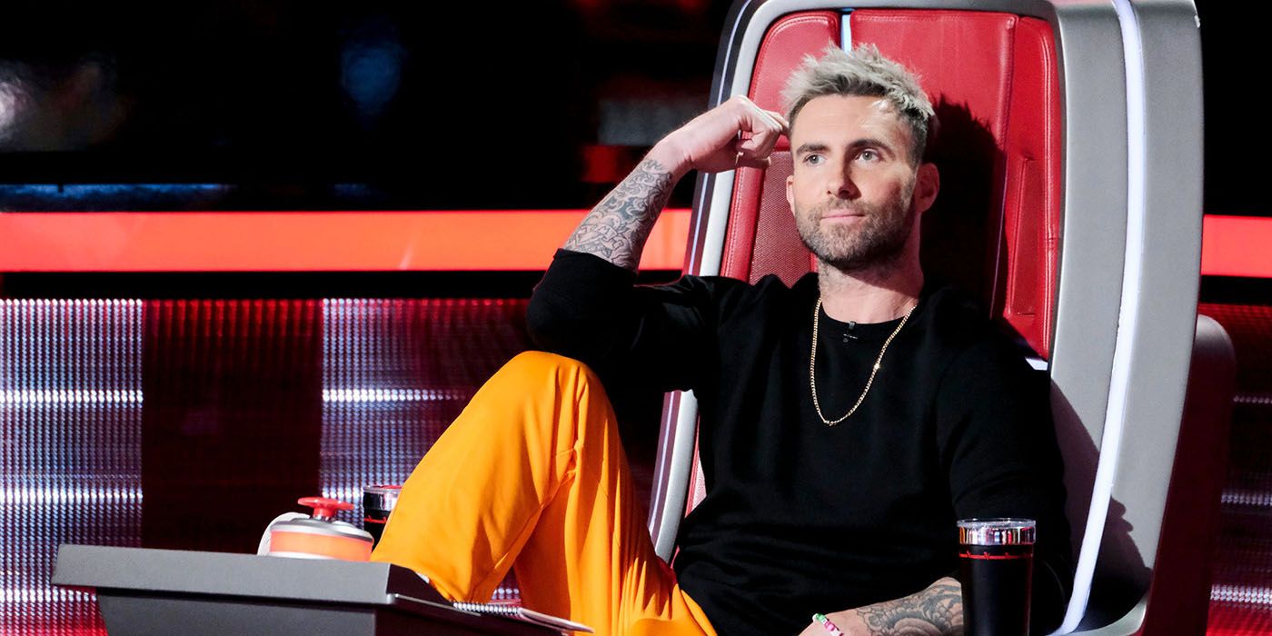 Adam Levine sitting in his chair on The Voice, knee up and hand to his head.