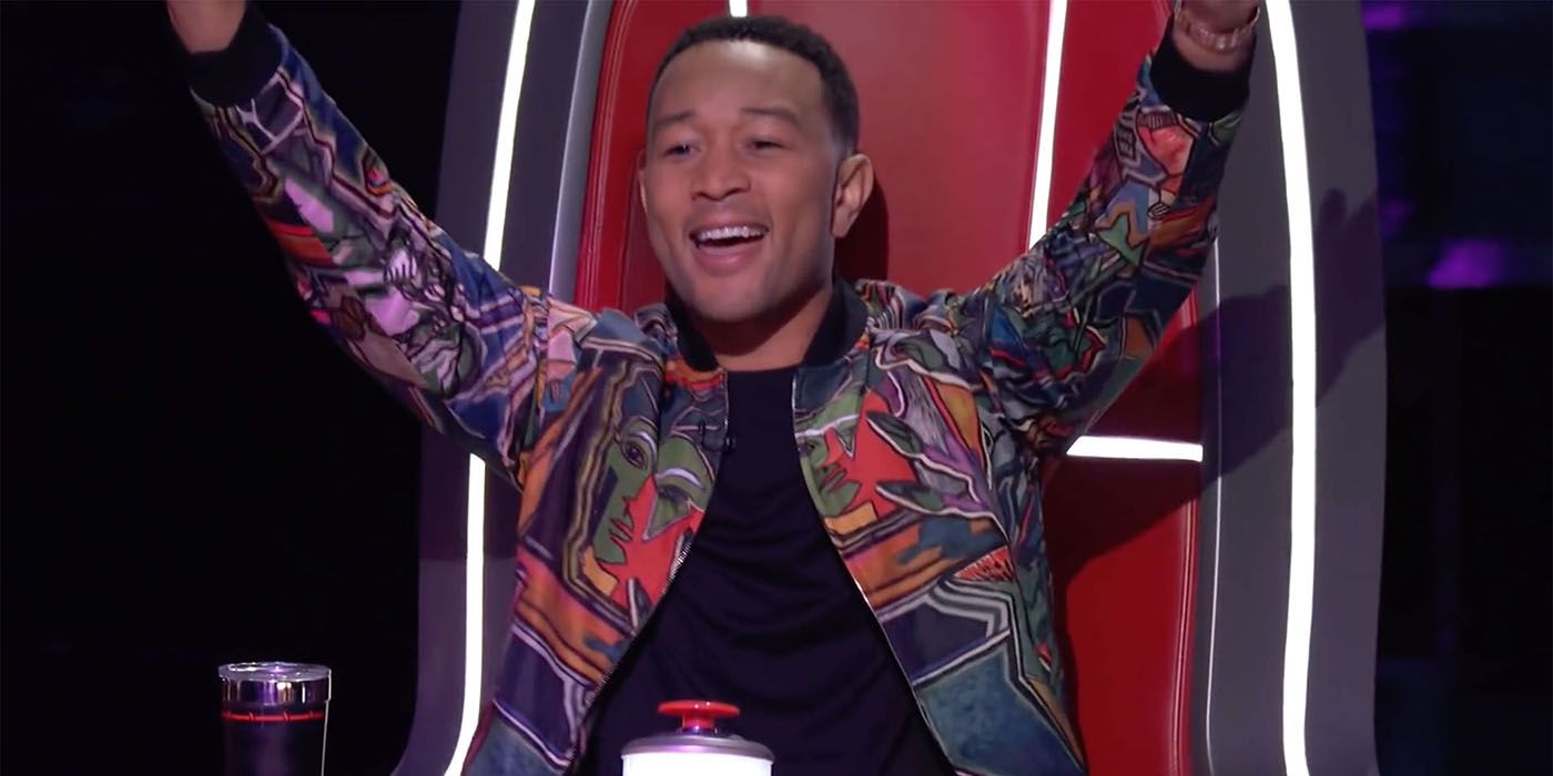 John Legend in his seat on The Voice, hands raised in the air.