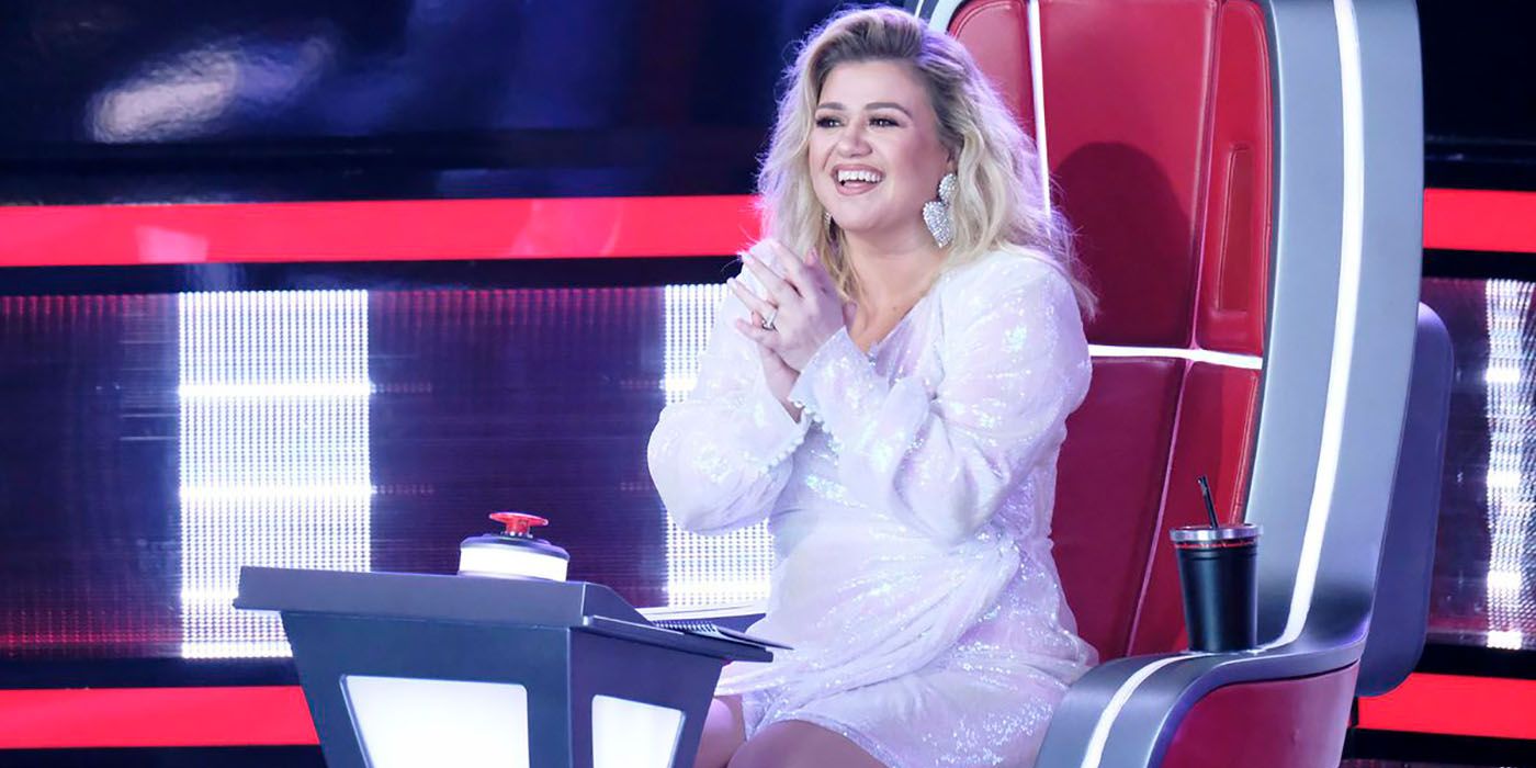 Kelly Clarkson sitting in her chair on The Voice, hands clasped in front of her, smile on her face.