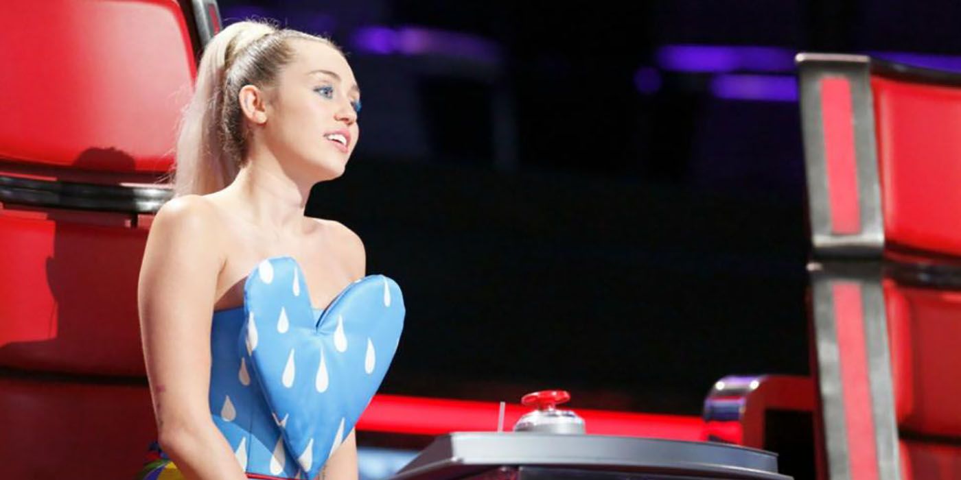 Miley Cyrus sitting in her chair on The Voice, wearing a shirt shaped like a big heart, head to the side.