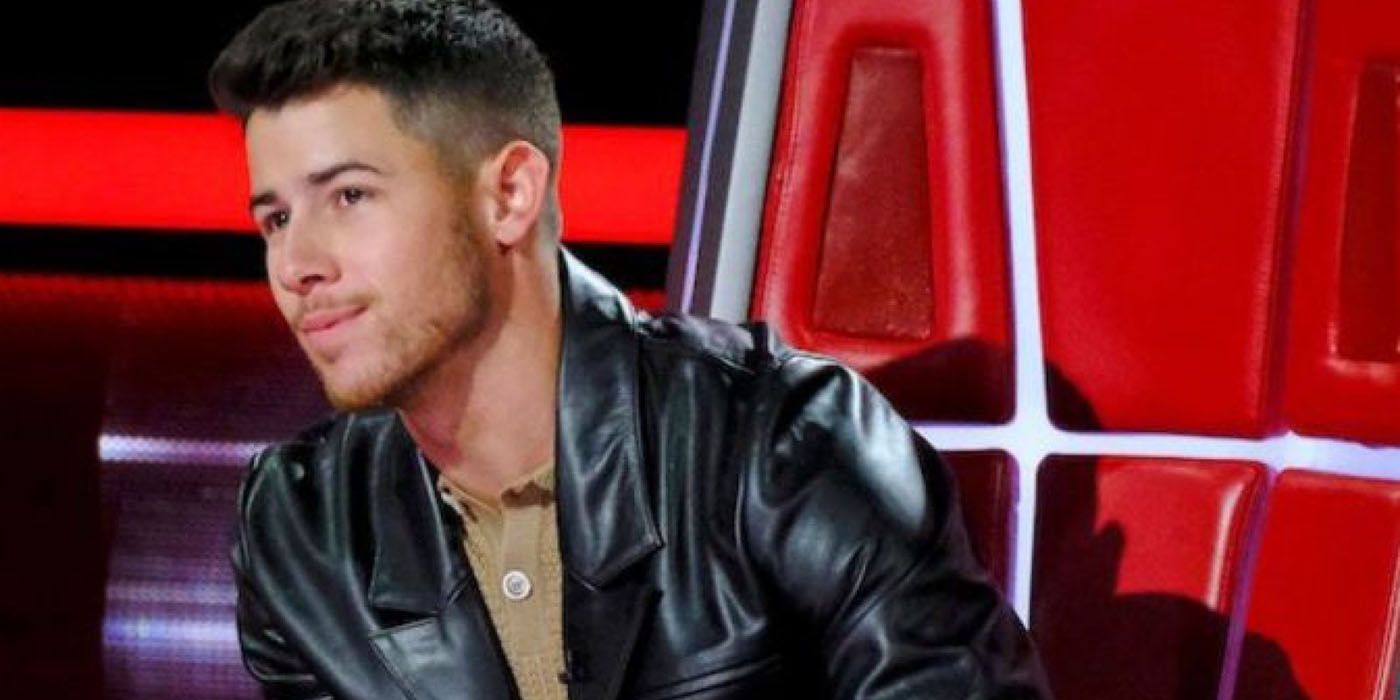 Nick Jonas in his chair on The Voice, leaning forward with a black leather jacket.