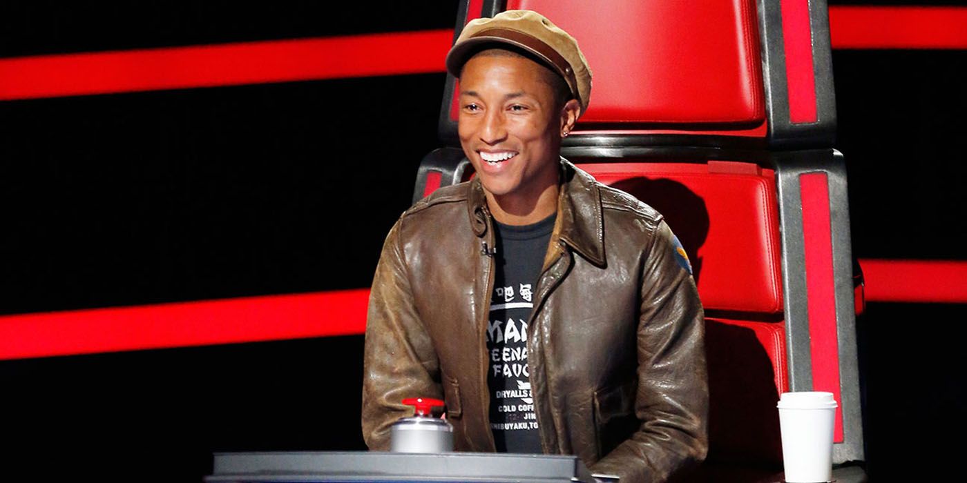 Pharrell Williams sitting in his chair on The Voice, leaning forward and smiling widely.