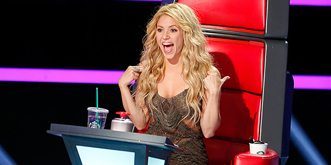 Shakira sitting in her chair on The Voice, hands on her shoulders, wide-mouthed.