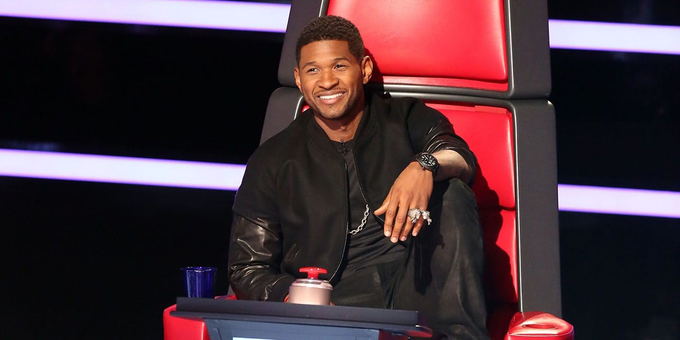 Usher sitting in his chair on The Voice, knee up with his arm draped over it.