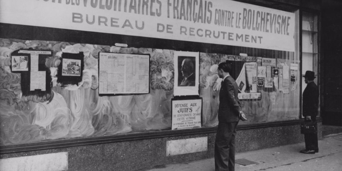 Two men stand in front of a wall that is covered in posters in French from The Sorrow and the Pity