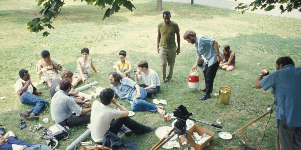 Various people sit on the grass while they are being filmed in Symbiopsychotaxiplasm