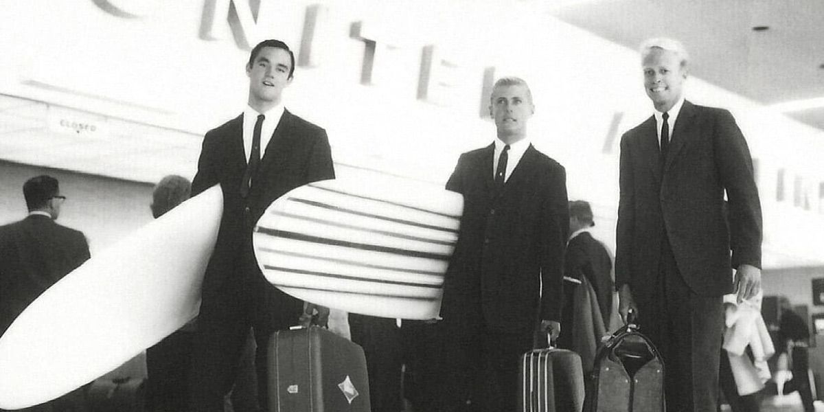 Three men in suits hold surfboards at the airport from The Endless Summer