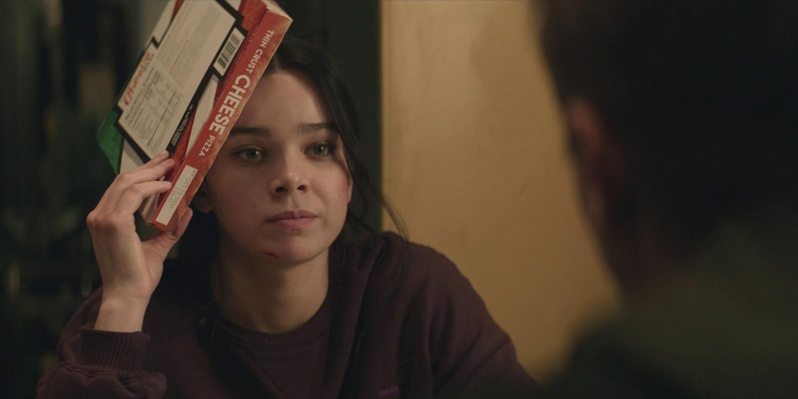 Kate Bishop wearing a maroon sweatshirt while holding a frozen pizza to her head in Hawkeye