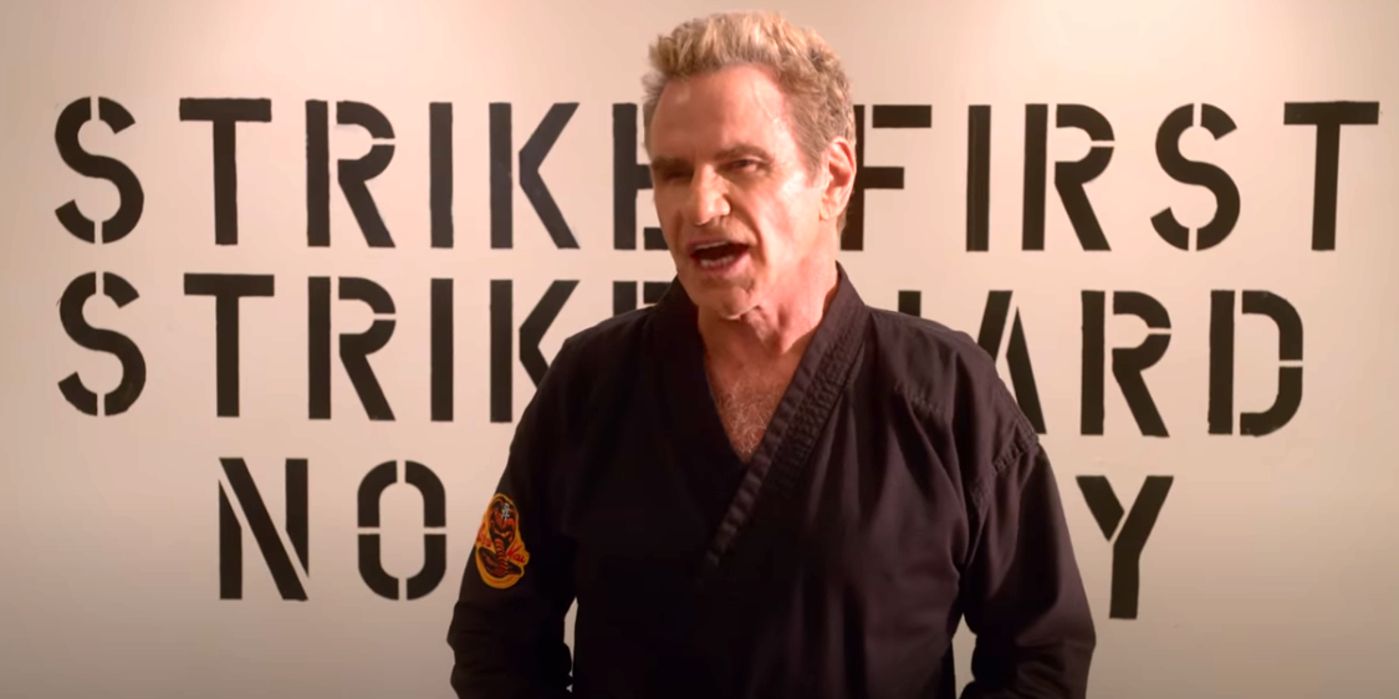 John Kreese standing in his gi by the Cobra Kai mantra sign.