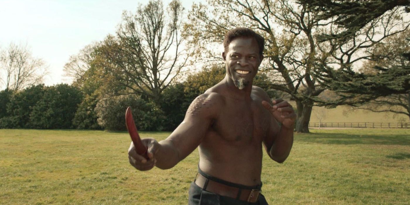 A shirtless Shola smiles while holding a knife in The King's Man